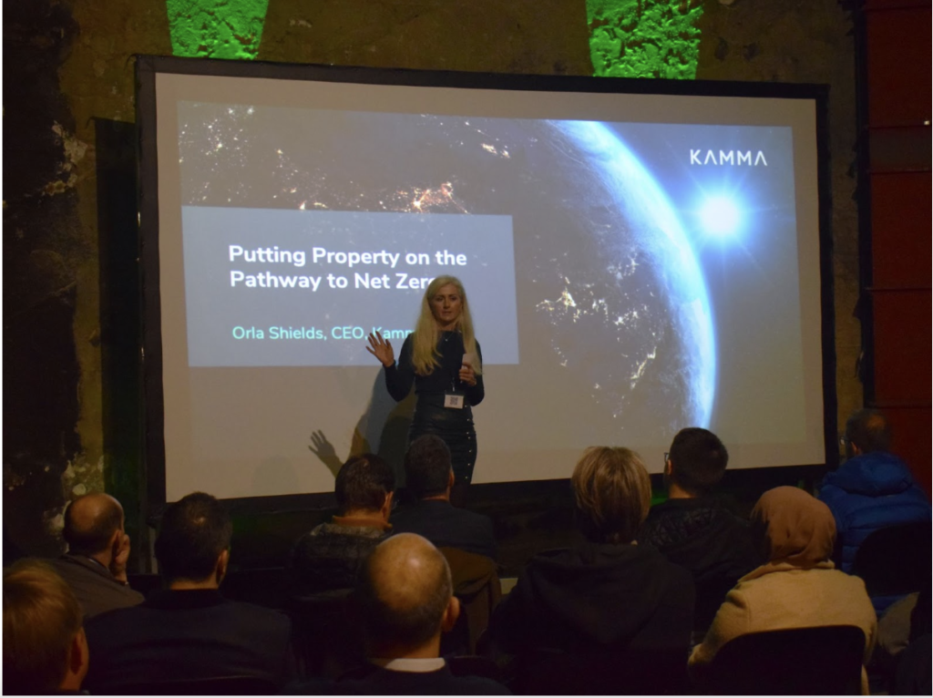 Kamma's CEO Orla Shields stands in front of a large screen which has the opening slide of the event presentation open, reading: 'Putting Property on the Path to Net Zero' 